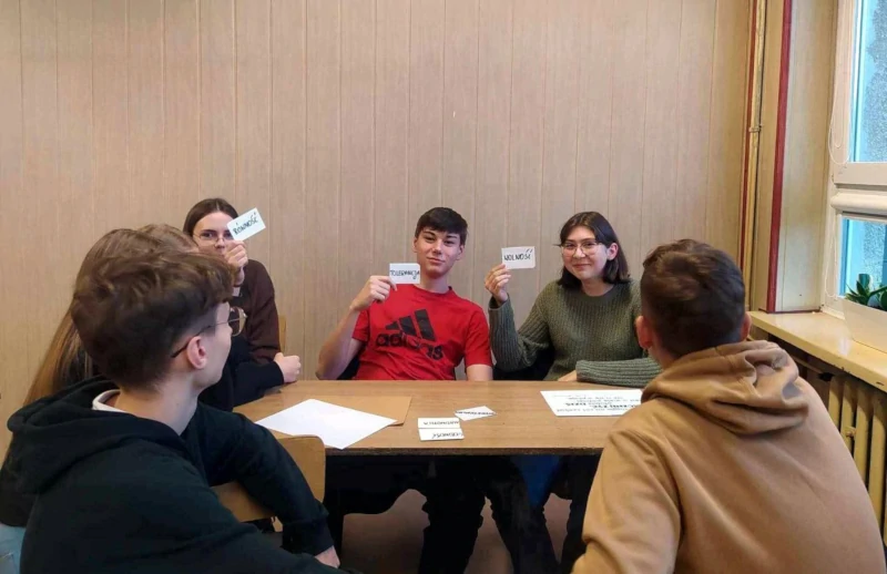 Uczniowie siedzący przy stolikach i pokazujący karteczki ze słowami