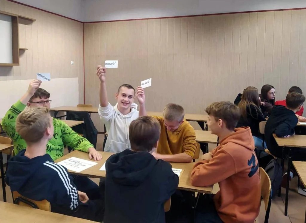 Uczniowie siedzący przy stolikach i pokazujący karteczki ze słowami
