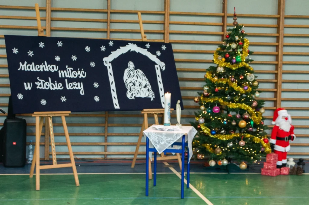 Dekoracje na akademii bożonarodzeniowej: tablica z napisem oraz szopką, choinka z mikołajem, stolik ze świecą i świętą figurką