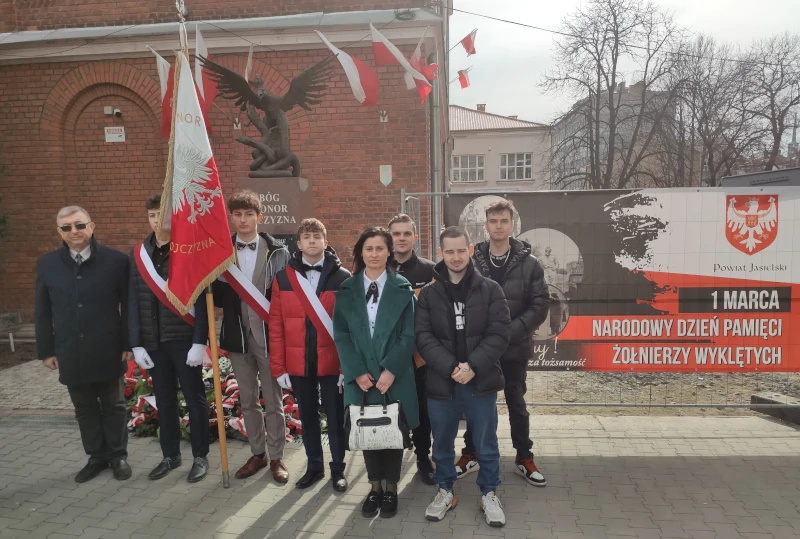 Dyrektor ZST w Jaśle wraz z pocztem sztandarowym, uczniami oraz nauczycielką na tle Banera z napisem "1 marca Narodowy Dzień Pamięci Żołnierzy Wyklętych"